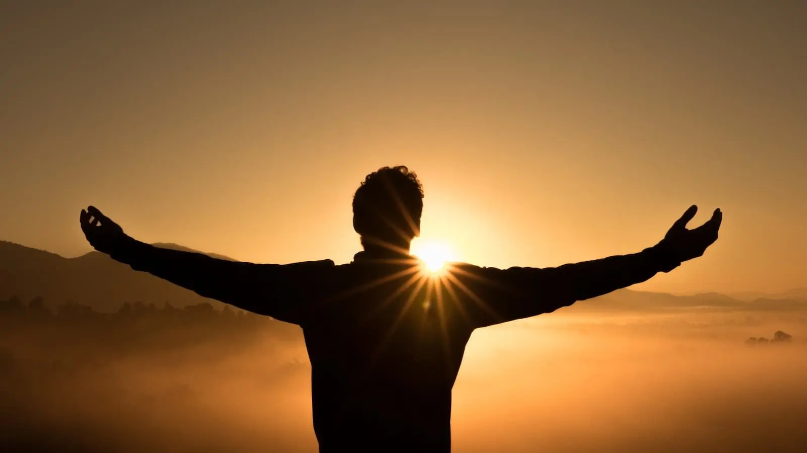A silhouette of a man with his arms outstretched in front of the sun.
